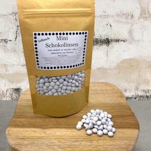 Könnte beinhalten: Eine Tüte mit weißen Schokoladen-Mini-Backstückchen, beschriftet mit "Vollmich Mini Schokolinsen" und dem Text "Zum einfach so naschen oder dekorieren von Kuchen. 400 Gramm." Ein kleiner Haufen weißer Schokoladenstückchen liegt auf einer Holzoberfläche.