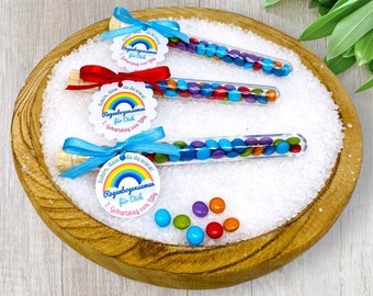 Op de afbeelding: Drie doorzichtige glazen buisjes gevuld met kleurrijke snoepjes. Elke buis is vastgebonden met een blauw lint en een rond label met een regenboogontwerp en de tekst "Schön, dass du da warst! Regenbogensamen für Dich 7. Geburtstag von Lilly".