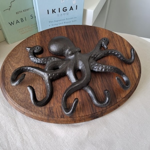 May include: A dark brown octopus wall hook mounted on a wooden oval plaque. The octopus is dark grey and has eight curved tentacles. The plaque is made of dark wood. The background includes books with the titles "Wabi Sabi" and "Ikigai".