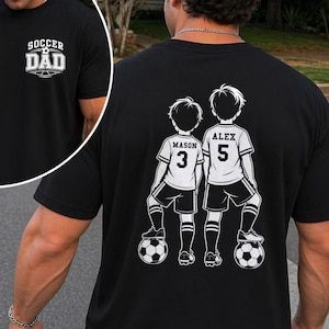 May include: Black t-shirt with the text "SOCCER DAD" on the front. The back of the shirt features a graphic of two soccer players with the names "MASON" and "ALEX" and their jersey numbers, 3 and 5, respectively. The players are standing on soccer balls.