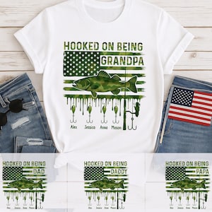 May include: White t-shirt with a camouflage American flag design and the text "HOOKED ON BEING GRANDPA". A green fish graphic and fishing hooks are also featured. The names Alex, Jessica, Anna, and Mason are listed below the hooks.