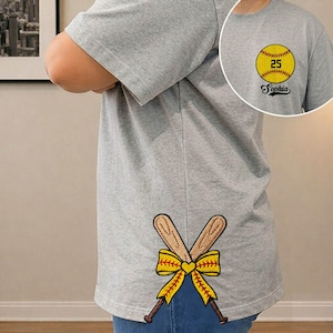 May include: Gray t-shirt with a softball graphic on the front and crossed baseball bats with a yellow bow on the back. The softball graphic includes the number 25 and the name "Sophia".