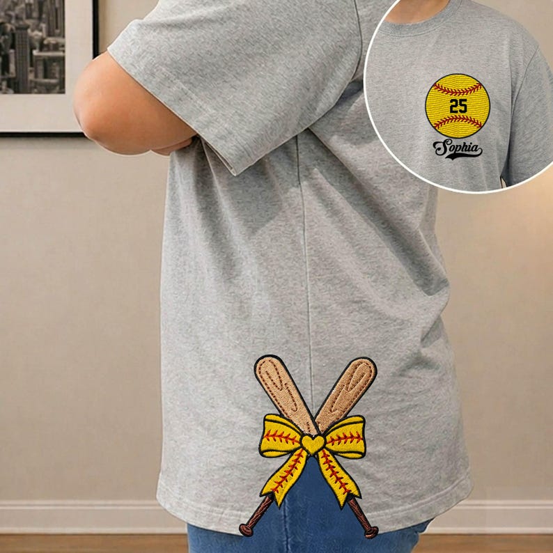 May include: Gray t-shirt with a softball graphic on the front and crossed baseball bats with a yellow bow on the back. The softball graphic includes the number 25 and the name "Sophia".