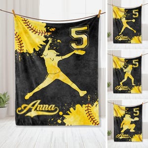 May include: Black and yellow softball themed fleece blanket with a silhouette of a player throwing a ball. The blanket has the number "5" and the name "Anna" printed on it.
