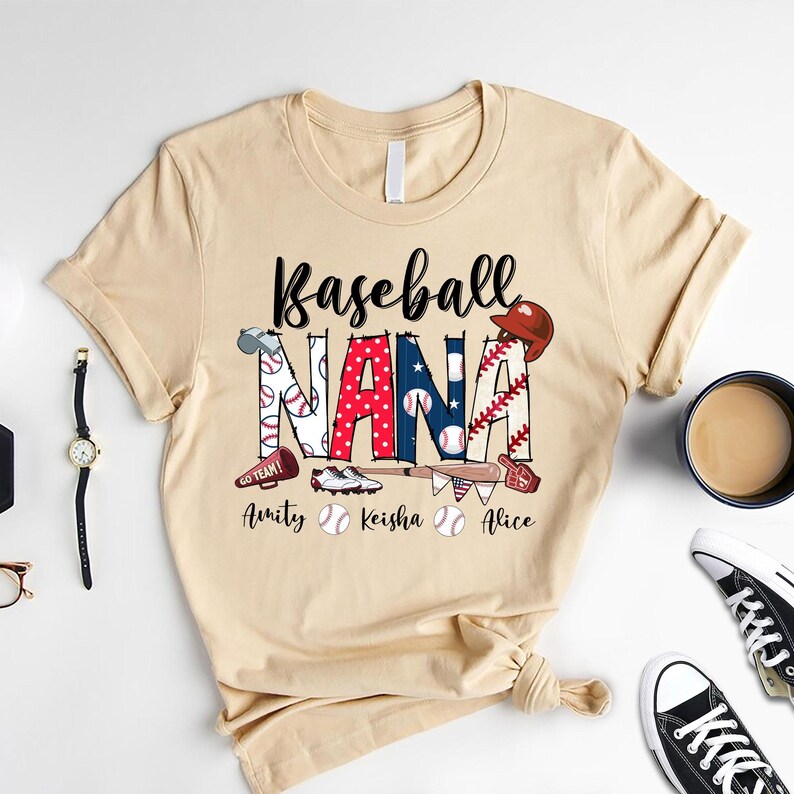 Puede incluir: Una camiseta beige con el texto "Baseball Nana" en letras negras. El texto est&aacute; rodeado de gr&aacute;ficos de tem&aacute;tica de b&eacute;isbol, incluyendo bates, pelotas y un guante. Los nombres "Amity", "Keisha" y "Alice" est&aacute;n impresos debajo del texto principal.