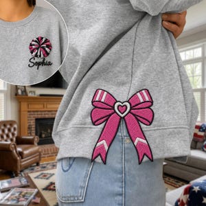 May include: A heather gray sweatshirt featuring a large embroidered pink bow with white accents on the lower side. The upper left chest area displays an embroidered pink and black pom-pom graphic above the black embroidered text Sophia.