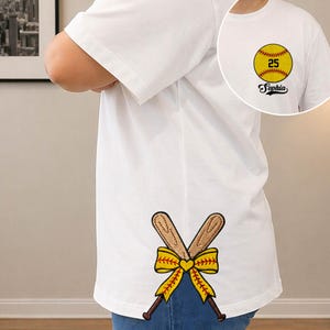 May include: White t-shirt with a softball design. The front features a yellow softball with the number 25 and the name "Sophia". The back has crossed baseball bats with a yellow bow and heart.