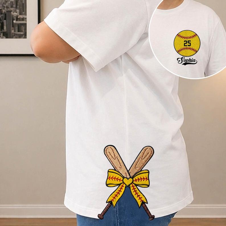 May include: White t-shirt with a softball design. The front features a yellow softball with the number 25 and the name "Sophia". The back has crossed baseball bats with a yellow bow and heart.