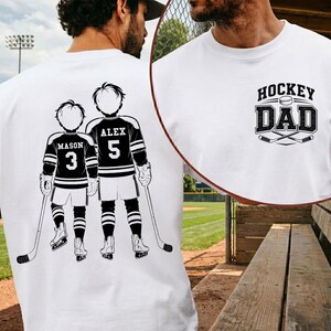 May include: White t-shirt with a hockey-themed design. The back features a graphic of two hockey players with the names "Mason" and "Alex" and the numbers "3" and "5". The front has the text "Hockey Dad" with crossed hockey sticks.