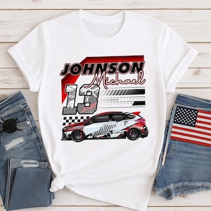 May include: White t-shirt with a racing car design, the name "JOHNSON Michael", and the number "13". The car is red, white, and black. The shirt is displayed with denim jeans and a small Union Jack flag.