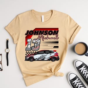 May include: Beige t-shirt with a race car design. The design includes the name "Johnson Michael" and the number "13". The car is white, red, and black. The shirt has a crew neck and short sleeves.