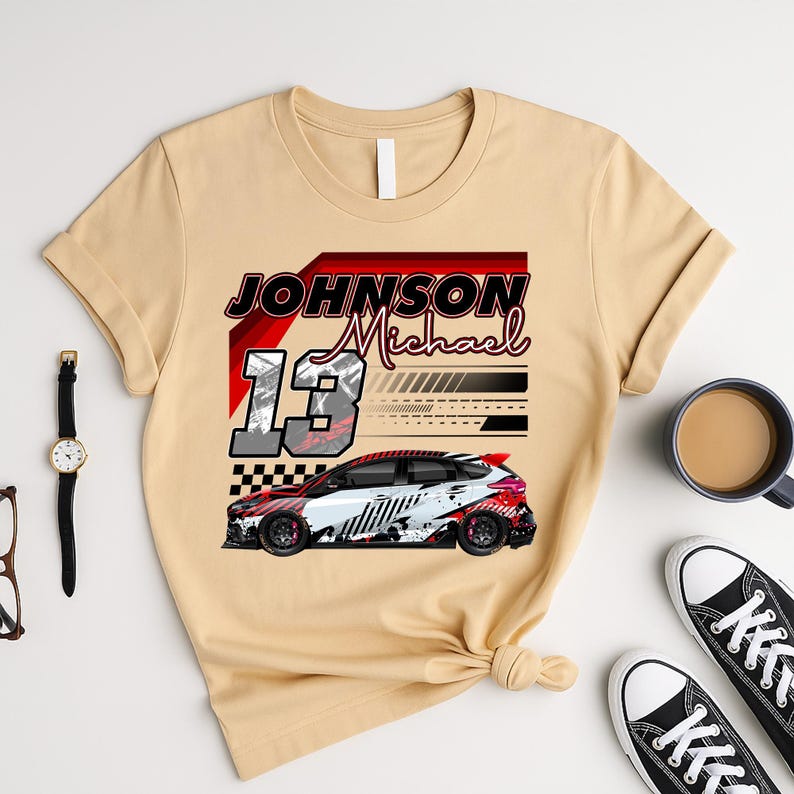 May include: Beige t-shirt with a race car design. The design includes the name "Johnson Michael" and the number "13". The car is white, red, and black. The shirt has a crew neck and short sleeves.