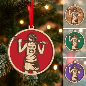 May include: Wooden Christmas ornament featuring a hockey player silhouette with the name "Sophia" and the number "25". The ornament has a red background and a natural wood border, with a red ribbon for hanging. Additional ornaments with green, brown, and purple backgrounds are also shown.