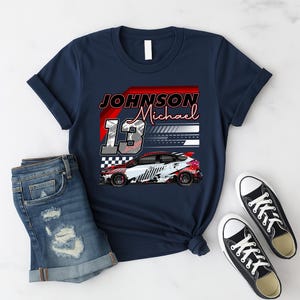 May include: Navy blue t-shirt with a race car graphic and the text "JOHNSON Michael 13". The design includes a red and white race car with black wheels. The shirt is paired with denim shorts and black and white trainers.