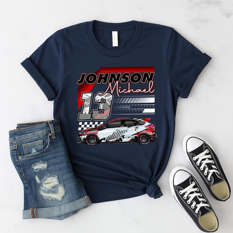 May include: Navy blue t-shirt with a race car graphic and the text "JOHNSON Michael 13". The design includes a red and white race car with black wheels. The shirt is paired with denim shorts and black and white trainers.