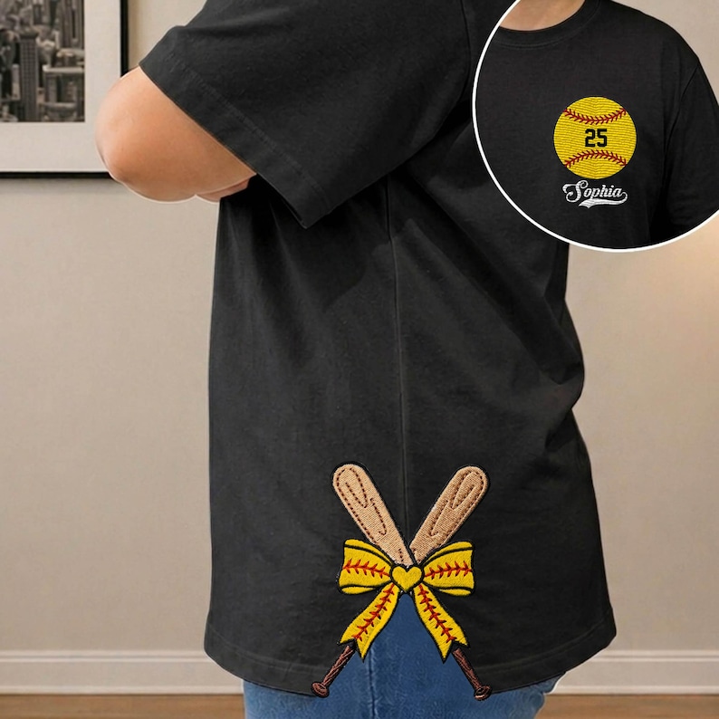 May include: Black t-shirt featuring a yellow softball with the number 25 and the name "Sophia". The shirt also has an embroidered design of crossed baseball bats and a yellow bow.
