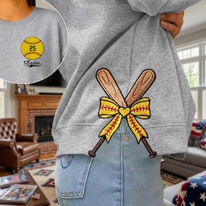 May include: Gray sweatshirt with a softball graphic and the name "Sophia". The back of the sweatshirt features crossed baseball bats with a yellow bow. The softball graphic includes the number "25".