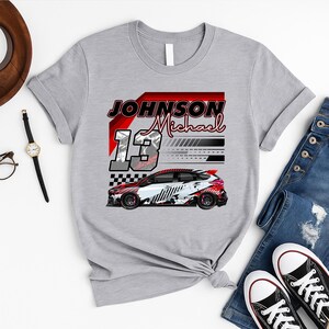 May include: A grey t-shirt with a race car graphic, the name "Johnson Michael", and the number "13". The car design includes red, white, and black details, and a checkered flag pattern.