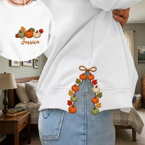 May include: A white sweatshirt featuring an embroidered design of orange, green, and cream pumpkins with autumn leaves. The chest area displays the pumpkins with the name Jessica in orange script, while the hem features a hanging pumpkin garland.