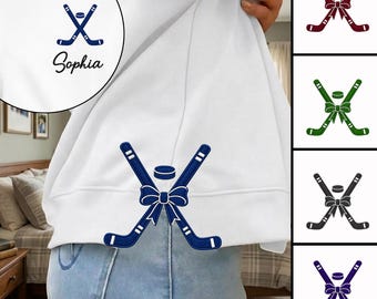 May include: A white sweatshirt featuring a dark blue embroidered graphic of two crossed hockey sticks tied with a bow and a hockey puck above. A circular inset shows the same graphic with the name Sophia printed in black script below it.