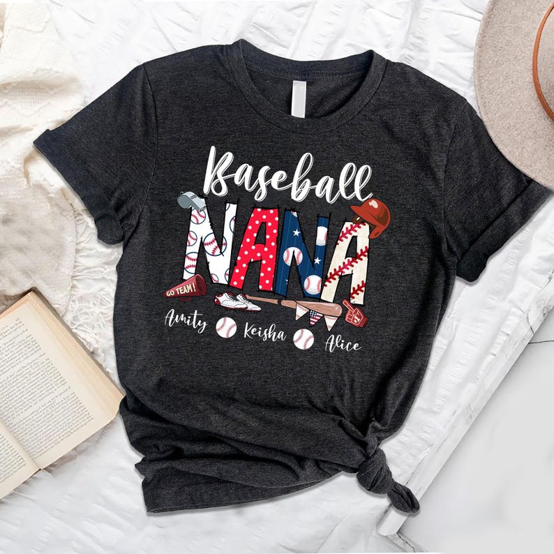 Puede incluir: Una camiseta gris oscuro con el texto "Baseball Nana" en letras blancas. Las letras est&aacute;n decoradas con gr&aacute;ficos de tem&aacute;tica de b&eacute;isbol, incluyendo pelotas de b&eacute;isbol, bates y un guante. Los nombres "Amity", "Keisha" y "Alice" est&aacute;n impresos debajo del texto principal.