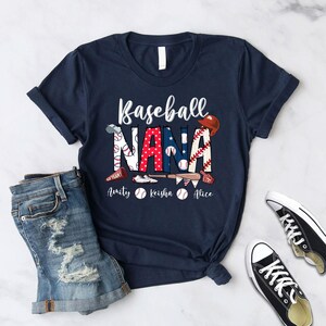 Puede incluir: Una camiseta azul marino con el texto "Baseball Nana" en letras blancas. El texto est&aacute; decorado con gr&aacute;ficos de tem&aacute;tica de b&eacute;isbol, incluyendo una gorra de b&eacute;isbol, bates y pelotas de b&eacute;isbol. Los nombres "Amity", "Keisha" y "Alice" est&aacute;n impresos debajo del texto principal.