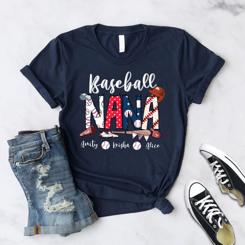 Puede incluir: Una camiseta azul marino con el texto "Baseball Nana" en letras blancas. El texto est&aacute; decorado con gr&aacute;ficos de tem&aacute;tica de b&eacute;isbol, incluyendo una gorra de b&eacute;isbol, bates y pelotas de b&eacute;isbol. Los nombres "Amity", "Keisha" y "Alice" est&aacute;n impresos debajo del texto principal.
