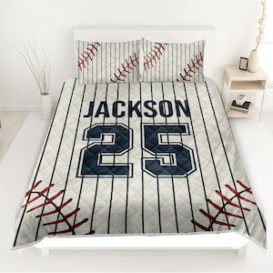 May include: A baseball-themed bedding set featuring a comforter and two pillowcases. The set has a cream background with vertical black stripes and baseball stitching details. The comforter displays the name "JACKSON" and the number "25" in navy blue.