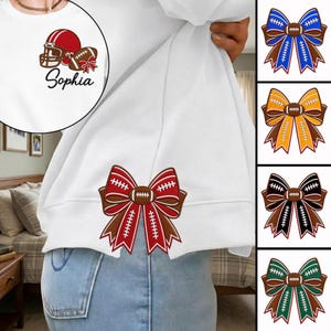 May include: A white sweatshirt featuring a red football helmet and a brown football with a red bow graphic, accompanied by the name Sophia. The sweatshirt also displays a large brown and red football-themed bow graphic on the lower hem.