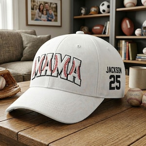 May include: A white baseball cap with the word "MAMA" in black outlined letters with red baseball stitching. The name "JACKSON" and the number "25" are on the side of the cap. The cap is sitting on a wooden table.