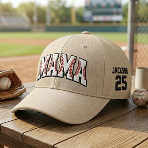 以下が含まれることがあります： ベージュ色の野球帽。白で「MAMA」の文字が刺繍され、赤い野球のステッチが施されています。「Jackson」という名前と「25」の数字が側面にあります。野球ボールとグローブが背景にあります。