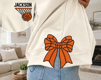 Puede incluir: Una sudadera color crema con un gráfico grande de un lazo naranja con diseño de baloncesto en la parte inferior de la espalda y un gráfico más pequeño en el pecho izquierdo que muestra una canasta de baloncesto y el nombre JACKSON.