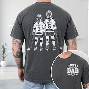May include: Dark gray t-shirt featuring a graphic of two hockey players with the names "Sophia" and "Anna" and numbers 3 and 5. The shirt also has a smaller graphic that says "Hockey Dad".