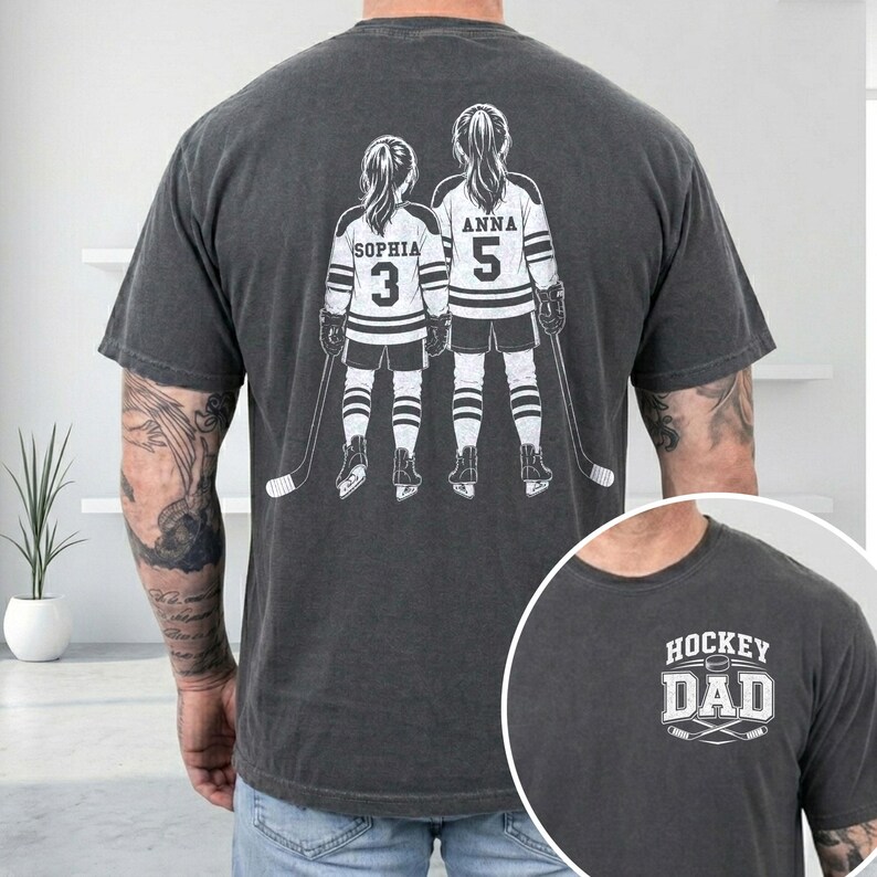May include: Dark gray t-shirt featuring a graphic of two hockey players with the names "Sophia" and "Anna" and numbers 3 and 5. The shirt also has a smaller graphic that says "Hockey Dad".