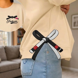 Peut inclure: Sweat-shirt beige avec un motif de ceinture noire et blanche dans le dos. Le devant pr&eacute;sente un motif de rouge &agrave; l&egrave;vres et de n&oelig;ud avec le nom "Jackson". Le sweat-shirt est port&eacute; avec un jean bleu.