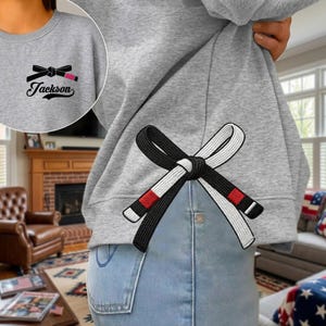 Peut inclure: Sweat-shirt gris avec un motif de ceinture de Jiu-Jitsu brod&eacute; en noir et blanc au dos. Le devant pr&eacute;sente une version plus petite du motif de la ceinture et le nom "Jackson". Le motif comprend une ceinture noire avec des rayures rouges.
