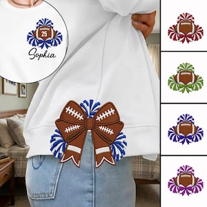 May include: A white sweatshirt featuring an embroidered design of a brown football with white laces, surrounded by blue pom-poms and a bow. The design includes the number 25 and the name Sophia. Additional color variations are shown on the right.
