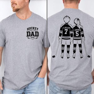 May include: A heather gray t-shirt with the text "HOCKEY DAD" and crossed hockey sticks. The back of the shirt features a black and white illustration of two hockey players with the names "LUCAS" and "ANNA" and their jersey numbers.