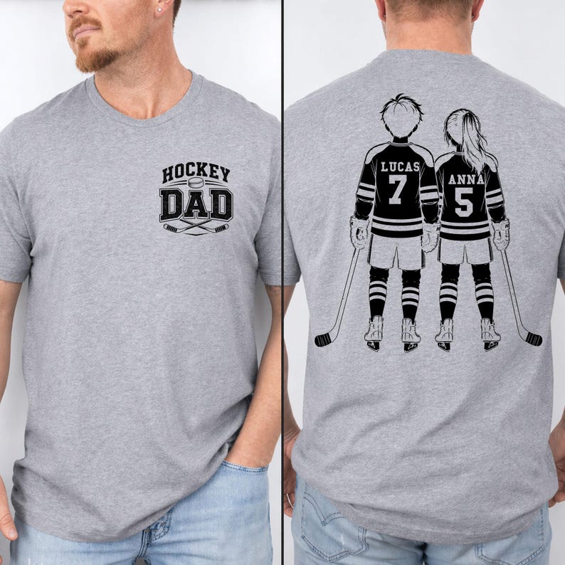 May include: A heather gray t-shirt with the text "HOCKEY DAD" and crossed hockey sticks. The back of the shirt features a black and white illustration of two hockey players with the names "LUCAS" and "ANNA" and their jersey numbers.