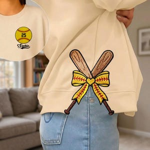 May include: Cream-colored sweatshirt with embroidered softball and crossed bats. The softball has the number 25 and the name "Sophia" embroidered on it. The crossed bats are brown with a yellow bow.