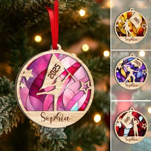 May include: Wooden Christmas ornament with a gymnast design. The ornament features a stained-glass effect with pink and purple hues, the year 2025, and the name "Sophia". It includes star cutouts and a red ribbon for hanging.
