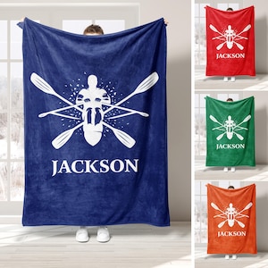 May include: A navy blue fleece blanket with a white rowing crew graphic and the name "JACKSON". Other blankets in red, green, and orange with the same design are displayed. The blankets are suitable for a bed or sofa.