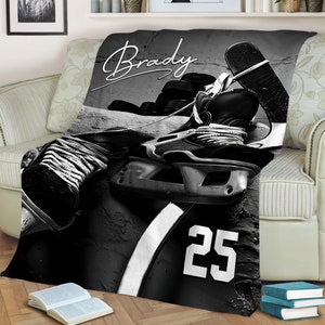 May include: Black and white blanket featuring hockey equipment, including skates, pucks, and a jersey with the number 25. The name "Brady" is written in a cursive font across the top. The blanket is draped over a sofa.