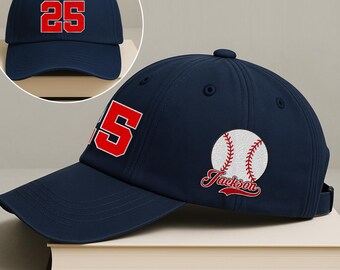 May include: A navy blue baseball cap with the number "25" in red on the front. The side of the cap features a baseball graphic with the name "Jackson" in red script. The cap is sitting on a book.