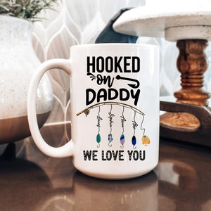 May include: White ceramic mug with a black design that says "Hooked on Daddy" with a fishing rod and four fishing lures with names on them. The text "We Love You" is at the bottom of the mug.
