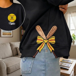 May include: Black sweatshirt with a softball and the name "Sophia" embroidered in white. The back features crossed baseball bats and a yellow bow with a heart in the center. The softball has the number "25".