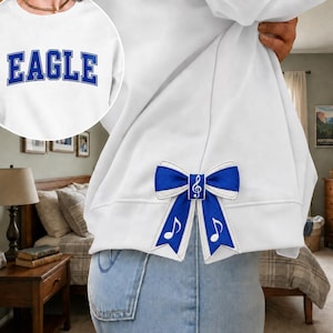 Puede incluir: Una sudadera blanca con la palabra EAGLE en letras azules de estilo universitario en el pecho y un lazo de tela azul y blanco sujeto al lateral, decorado con una clave de sol blanca y s&iacute;mbolos de notas musicales en las cintas.