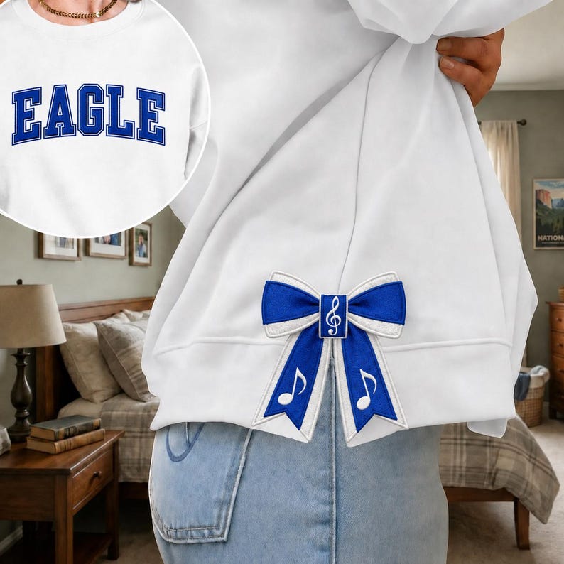 Puede incluir: Una sudadera blanca con la palabra EAGLE en letras azules de estilo universitario en el pecho y un lazo de tela azul y blanco sujeto al lateral, decorado con una clave de sol blanca y s&iacute;mbolos de notas musicales en las cintas.