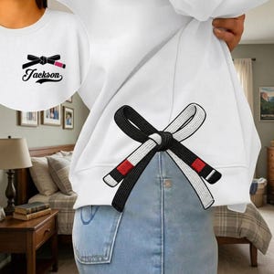 Peut inclure: Sweat-shirt blanc avec un motif de ceinture de Jiu-Jitsu noire et blanche brod&eacute; au dos. Le devant du sweat-shirt pr&eacute;sente une version plus petite du motif avec le nom "Jackson" en dessous. La ceinture a des accents rouges.