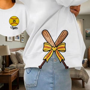 May include: White sweatshirt with embroidered softball and crossed bats design. The softball has the number 25 and the name "Sophia" below it. The crossed bats are brown with a yellow bow.